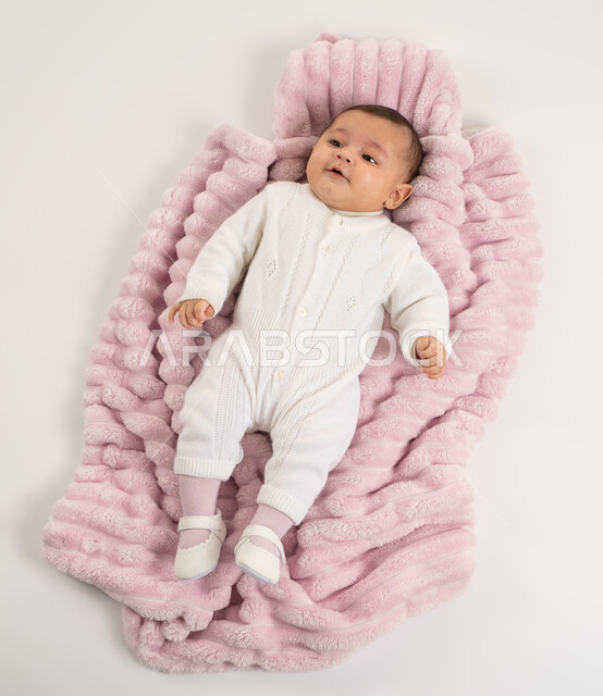 ايماءات اللعب والنظر إلى شيء ما, بورتريه لطفلة عربيه خليجيه سعودية لطيفة ترتدي بربتوز أبيض صوف مستلقية على غطاء فرو باللون الوردي, الاهتمام بصحة الأبناء والعناية بهم, خلفية بيضاء