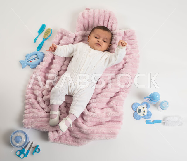 الاهتمام بصحة الأبناء والعناية بهم, مفهوم البراءة والحنان , أدوات الرضاعه ومستلزمات الرضيع, بورتريه لطفلة عربيه خليجيه سعودية لطيفة ترتدي بربتوز أبيض صوف مستلقية على غطاء فرو باللون الوردي, خلفية بيضاء
