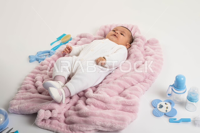 مفهوم البراءة والحنان , أدوات الرضاعه ومستلزمات الرضيع, بورتريه لطفلة عربيه خليجيه سعودية لطيفة ترتدي بربتوز أبيض صوف مستلقية على غطاء فرو باللون الوردي, الاهتمام بصحة الأبناء والعناية بهم, خلفية بيضاء