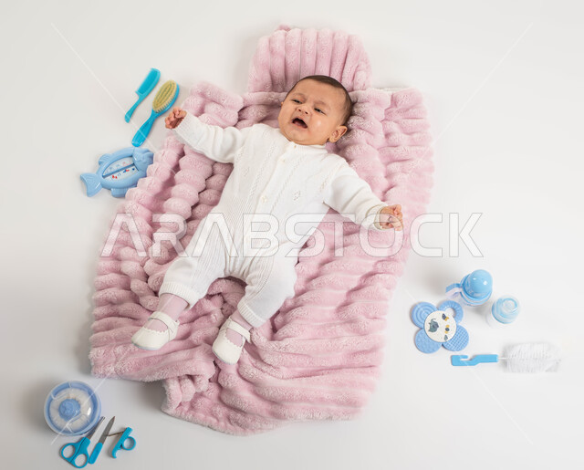 ايماءات البكاء والحاجة إلى شيء ما, أدوات الرضاعه ومستلزمات الرضيع, بورتريه لطفلة عربيه خليجيه سعودية لطيفة ترتدي لباس أبيض صوف مستلقية على غطاء فرو باللون الوردي, الاهتمام بصحة الأبناء والعناية بهم, خلفية بيضاء