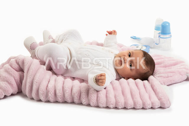 أدوات الرضاعه ومستلزمات الرضيع, الاهتمام بصحة الأبناء والعناية بهم, مفهوم البراءة والحنان, بورتريه لطفلة عربيه خليجيه سعودية لطيفة ترتدي بربتوز أبيض صوف مستلقية على غطاء فرو باللون الوردي, خلفية بيضاء