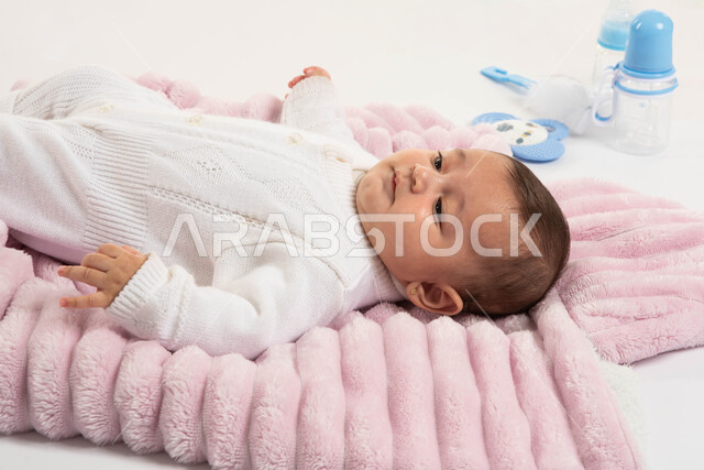 الاهتمام بصحة الأبناء والعناية بهم, أدوات الرضاعه ومستلزمات الرضيع, مفهوم البراءة والحنان, بورتريه مقرب لطفلة عربيه خليجيه سعودية لطيفة ترتدي بربتوز أبيض صوف مستلقية على غطاء فرو باللون الوردي, خلفية بيضاء
