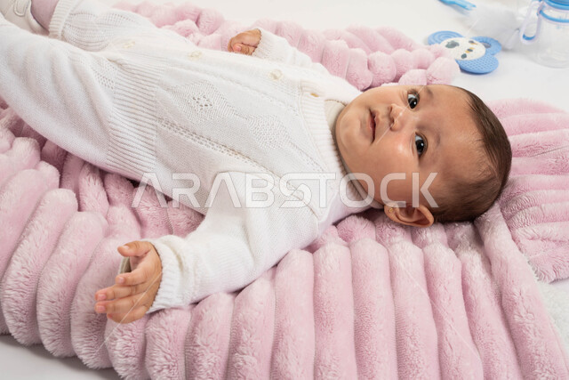 نظرات بريئة تملؤها اللطافة، بورتريه مقرب لطفلة عربية خليجية سعودية ترتدي بربتوز أبيض صوف تنظر إلى الكاميرا، إيماءات الحب والأمان، الاهتمام والعناية بالأطفال، مفهوم الأمومة والطفولة، خلفية بيضاء