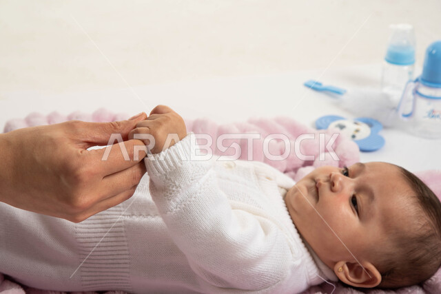 أم تمسك يد طفلتها، اللعب مع الأبناء وقضاء وقت ممتع معهم، حنان الأمهات والعناية بالأطفال، بورتريه مقرب لطفلة عربية خليجية سعودية ترتدي بربتوز أبيض صوف مستلقية تنظر إلى والدتها بإيماءات الحب والأمان، مفهوم الأمومة والطفولة، خلفية بيضاء