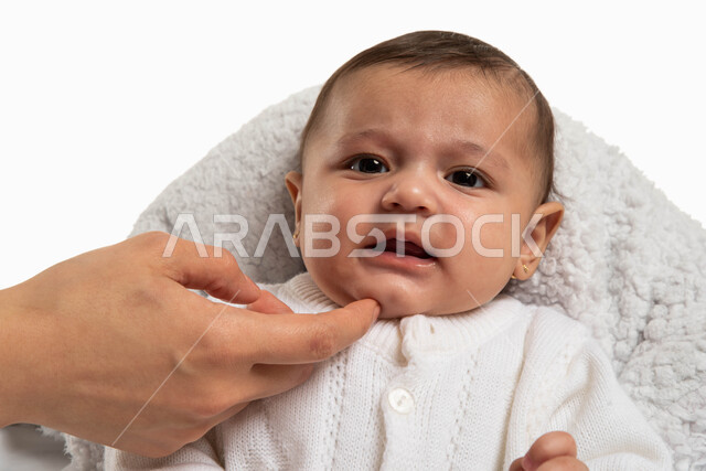 الاستمتاع بقضاء وقت ممتع مع الأطفال الرضع، حنان الأمهات والعناية بالأبناء، بورتريه مقرب لطفلة رضيعة تجلس في كرسي الطعام ترتدي بربتوز أبيض صوف, أم عربية خليجية سعودية تداعب طفلتها بلطف وحنان، مفهوم الأمومة والطفولة، خلفية بيضاء