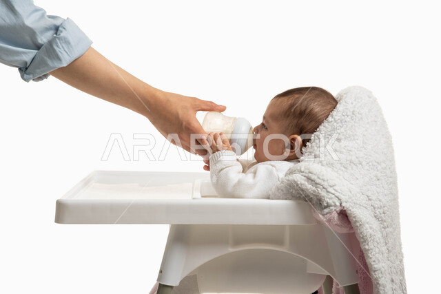 استخدام الحليب الصناعي المليء بالفيتامينات, الرعايه والاهتمام بتغذية ونمو الأبناء, أم عربية تقوم بإعطاء طفلتها الغذاء المفيد، بورتريه من الجانب  لطفلة عربية خليجية سعودية تقوم بشرب الحليب من عبوة الرضاعة تجلس على كرسي الأطفال، مشاعر العطف والحنان, خلفية بيضاء