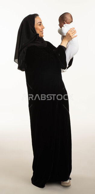 الأسرة السعودية وتحمل المسؤولية, الاستمتاع بقضاء وقت ممتع مع الأطفال, اللعب مع الأبناء بإيماءات السعادة، بورتريه مقرب لإمرأة عربية سعودية خليجية محجبة ترفع رضيعتها للأعلى، مفهوم الأمومة والطفولة، صورة الجسم بالطول الكامل, خلفية بيضاء