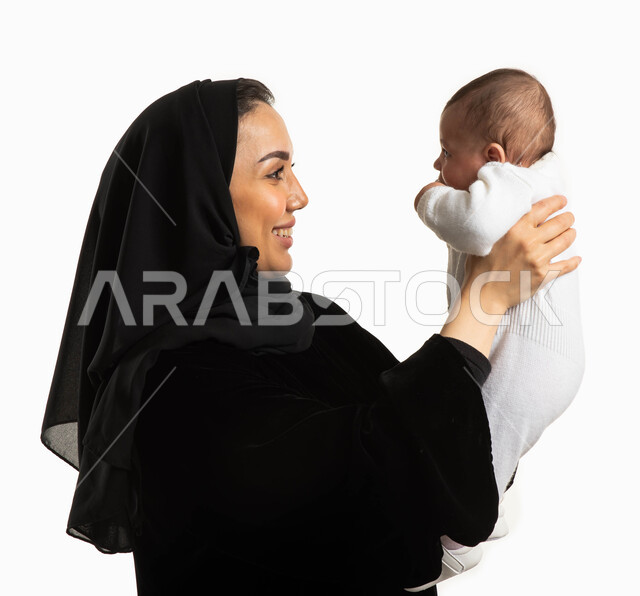 الاستمتاع بقضاء وقت ممتع مع الأطفال, اللعب مع الأبناء بإيماءات السعادة، بورتريه مقرب لإمرأة عربية سعودية خليجية  محجبة ترفع رضيعتها للأعلى، مفهوم الأمومة والطفولة، الأسرة السعودية وتحمل المسؤولية ، خلفية بيضاء