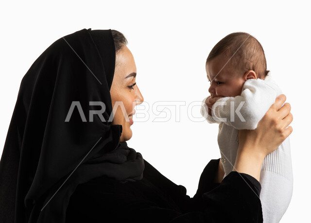 مداعبة الأم لأبنائها بإيماءات السعادة، حنان الأمهات و رعايتهم للأولاد، بورتريه مقرب لإمرأة عربية خليجية سعودية محجبة تحمل رضيعتها بحب، مفهوم  الأسرة السعودية وتحمل المسؤولية، الطمأنينة والشعور بالأمان، خلفية بيضاء