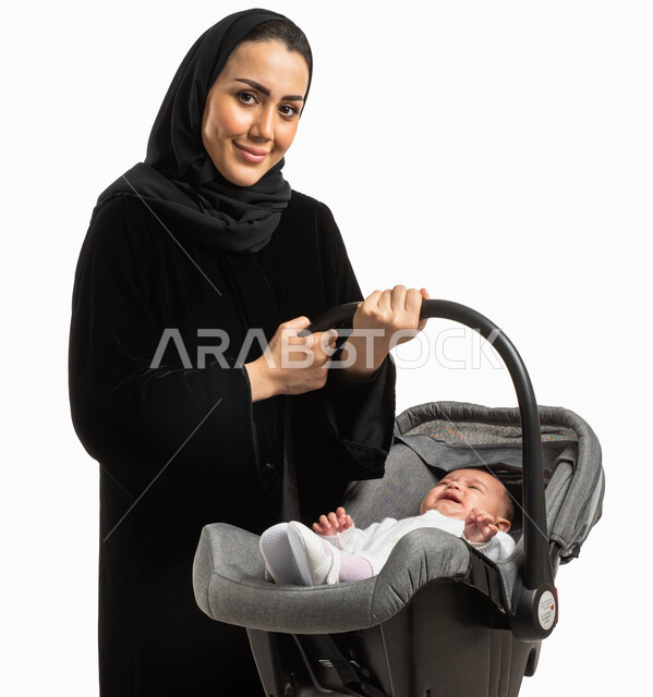 مفهوم الأمومة وتحمل المسؤولية والاهتمام بالأبناء, بورتريه لأم عربية خليجيه سعودية محجبة ترتدي العباءة السوداء مبتسمة تحمل كرسي طفلتها الصغيرة وتنظر إلى الكاميرا، إيماءات وجه تدل على البكاء والصراخ, خلفية بيضاء