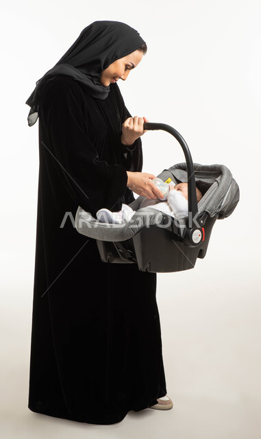 نظرات مليئة بالحب والحنان, شرب الحليب من عبوة الرضاعه, بورتريه  لأم عربية خليجيه سعودية محجبة ترتدي العباءة السوداء تحمل كرسي طفلتها الصغيرة، مفهوم الأمومة والرعايه والاهتمام بتغذية ونمو الأبناء, صورة الجسم بالطول الكامل, خلفية بيضاء