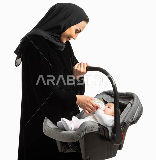 شرب الحليب من عبوة الرضاعه, بورتريه مقرب لأم عربية خليجيه سعودية محجبة مبتسمة ترتدي العباءة السوداء تحمل كرسي طفلتها الصغيرة، مفهوم الأمومة والرعايه والاهتمام بتغذية ونمو الأبناء, إيماءات وجه مليئة بالحنان, خلفية بيضاء