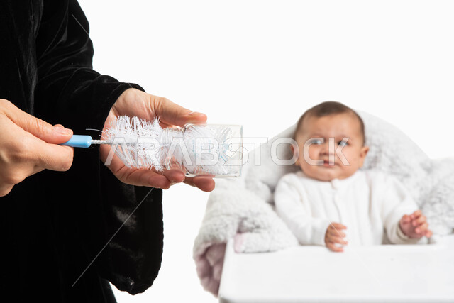 جلوس الرضيع على كرسي الطعام بإيماءات البكاء والإحساس بالجوع, الاعتناء بنظافة مستلزمات الطفل, بورتريه مقرب لأم عربية خليجيه سعودية تقوم بتنظيف وتعقيم عبوة الرضاعة باستخدام الفرشاة, الاهتمام بصحة الأبناء والوقاية من الأمراض والجراثيم, خلفية بيضاء