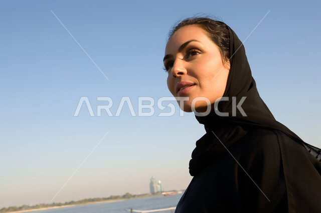 قضاء وقت العطلة على الشاطئ في دولة الإمارات المتحدة، صورة مقربة لإمرأة عربية خليجية إماراتية محجبة ترتدي العباءة السوداء تقوم بتأمل البحر، السياحة الساحلية الترفيهية المسلية، تامل أمواج البحار الهادئة في وضح النهار