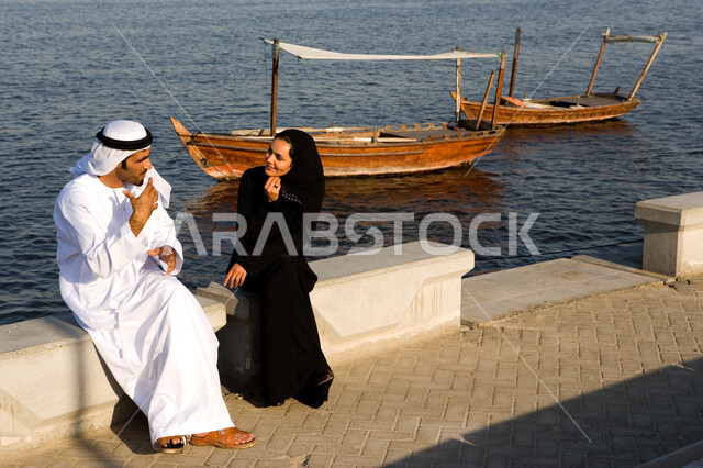 قضاء وقت ممتع في جلسات رومانسية على الكورنيش في وضح النهار، زوجان عربيان خليجيان إماراتيان يرتديان الزي الإماراتي التقليدي يقومان بتبادل الأحاديث أثناء الجلوس على شاطئ البحر في دولة الإمارات المتحدة، السياحة الساحلية الترفيهية المسلية