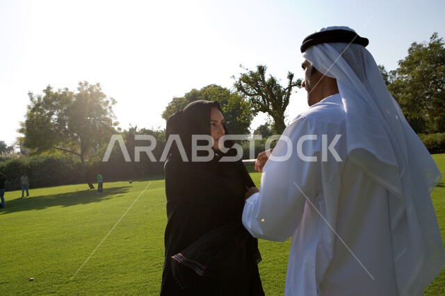 اختلاف وجهات النظر و التشاور في أمر ما، زوجان عربيان خليجيان اماراتيان يرتديان الزي الإماراتي التقليدي يقومان بالنقاش والحديث في الحديقة، ايماءات وجه ويدين تدل على الإختلاف بالرأي، منظر العشب والأشجار الخضراء في وضح النهار