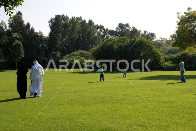 منظر العشب والأشجار الخضراء في وضح النهار، صورة من الخلف لزوجان عربيان خليجيان إماراتيان يرتديان الزي الإماراتي التقليدي يقومان بالمشي في الحديقة العامة في دولة الإمارات العربية المتحدة، أطفال يلعبون في المساحات الواسعة، صورة الجسم بالطول الكامل