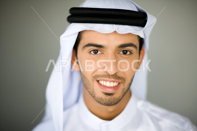 الاستمتاع بالتقاط صورة شخصية للوثائق الرسمية, الاهتمام بالمظهر الخارجي, بورتريه مقرب لرجل عربي خليجي إماراتي مبتسم يرتدي الكندورة والغترة ينظر إلى الكاميرا بإيماءت السرور والثقة, خلفية رمادية