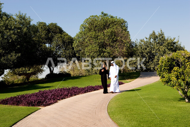 منظر العشب والأشجار الخضراء في وضح النهار، زوجان عربيان خليجيان إماراتيان يرتديان الزي الإماراتي التقليدي يتناقشان في موضوع ما يقومان بالمشي في الحديقة العامة في دولة الإمارات العربية، تبادل الأحايث ووجهات النظر، صورة الجسم بالطول الكامل