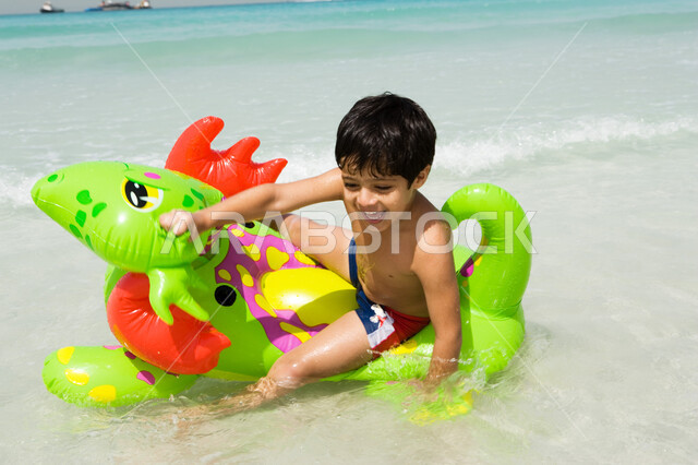 منتجعات شاطئية في الإمارات, صورة مقربة لصبي عربي إماراتي خليجي مبتسم يجلس على عوامة السباحة على شاطئ البحر, أنشطة ترفيهية مسلية في العطلة, إيماءات السعادة و الاستمتاع بالفعاليات الصيفية 