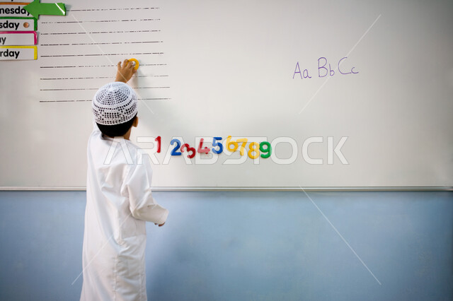 مدارس الامارات العربية المتحدة وأهداف التربية والتعليم، صورة من الخلف لتلميذ عربي خليجي اماراتي يرتدي الطاقية البيضاء والكندورة الاماراتية يقف أمام السبورة، العودة إلى المدارس والمقررات والمناهج المتبعة في الفصل، الأنشطة الحسابية وتنمية الذكاء لدى الأولاد