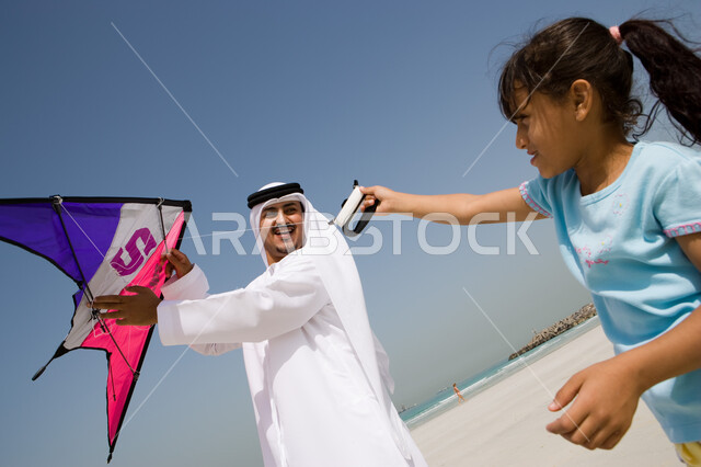 إيماءات السعادة والاستمتاع بقضاء الإجازة الصيفية مع الأبناء , أب عربي خليجي إماراتي يرتدي الكندورة والغترة الإماراتية يقوم باللعب مع ابنته بالطائرة الورقية, منتجعات شاطئية في الامارات , أنشطة ترفيهية مسلية في العطلة 