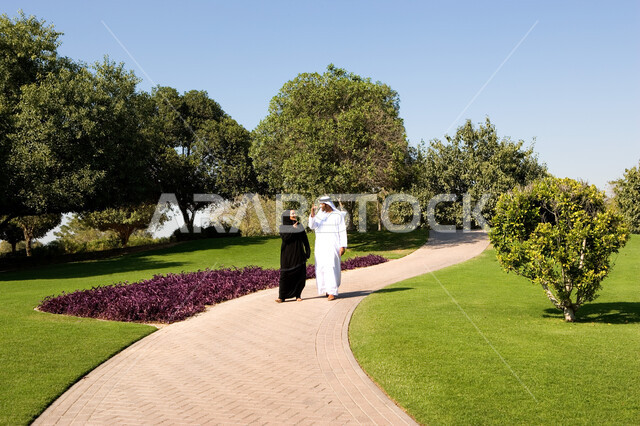 صورة الجسم بالطول الكامل، تبادل الأحايث ووجهات النظر، منظر العشب والأشجار الخضراء في وضح النهار، زوجان عربيان خليجيان إماراتيان سعيدان يرتديان الزي الإماراتي التقليدي يتناقشان في موضوع ما يقومان بالمشي في الحديقة العامة في دولة الإمارات العربية المتحدة