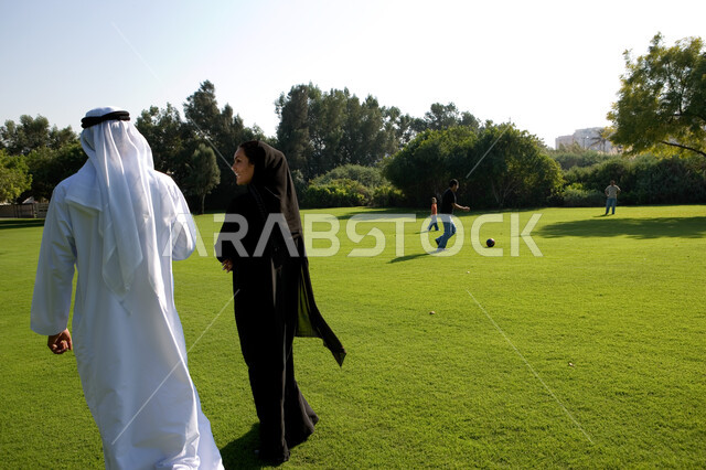 منظر العشب والأشجار الخضراء في وضح النهار، صورة من الخلف لزوجان عربيان خليجيان إماراتيان يرتديان الزي الإماراتي التقليدي يقومان بالمشي في الحديقة العامة في دولة الإمارات العربية المتحدة، أطفال يلعبون في المساحات الواسعة، صورة الجسم بالطول الكامل