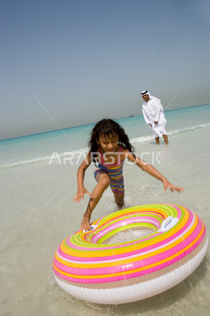 فتاة صغيرة تقوم باللعب بعوامة السباحة رفقة , إيماءات السعادة والاستمتاع بقضاء الإجازة الصيفية مع الأبناء , أب عربي خليجي إماراتي يرتدي الكندورة والغترة الإماراتية يقوم باللعب مع ابنته على الشاطئ, منتجعات شاطئية في الامارات , أنشطة ترفيهية مسلية في العطلة