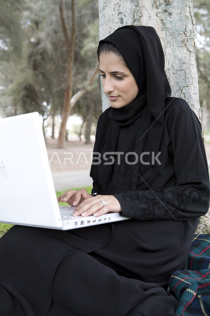 الجلوس في الهواء الطلق، إنجاز مهام العمل، احتراف وظائف الحاسوب وتكنولوجيا المعلومات، إمرأة عربية خليجية اماراتية محجبة تستخدم الحاسوب المحمول, إستخدام أجهزة تقنية حديثة، تصفح الإنترنت، خلفية الطبيعة الخضراء، جلسة عمل عن بعد