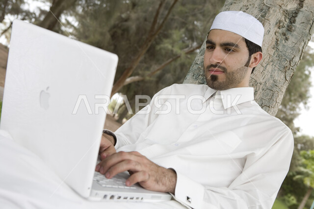 احتراف وظائف الحاسوب وتكنولوجيا المعلومات، إنجاز مهام العمل، رجل عربي خليجي سعودي يستخدم الحاسوب المحمول, إستخدام أجهزة تقنية حديثة، تصفح الإنترنت، الجلوس في الهواء الطلق، خلفية الطبيعة الخضراء، جلسة عمل عن بعد