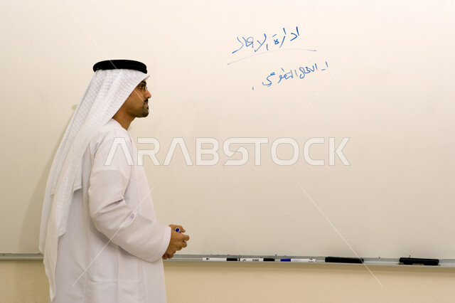 مدرس مختص في يقوم بشرح محاضرة عن إدارة الأعمال في الجامعة، صورة مقربة لأستاذ جامعي عربي خليجي إماراتي يرتدي الغترة والكندورة الإماراتية ويكتب النقاط الهامة والملاحظات على السبورة البيضاء، التعليم الجامعي في الإمارات العربية المتحدة