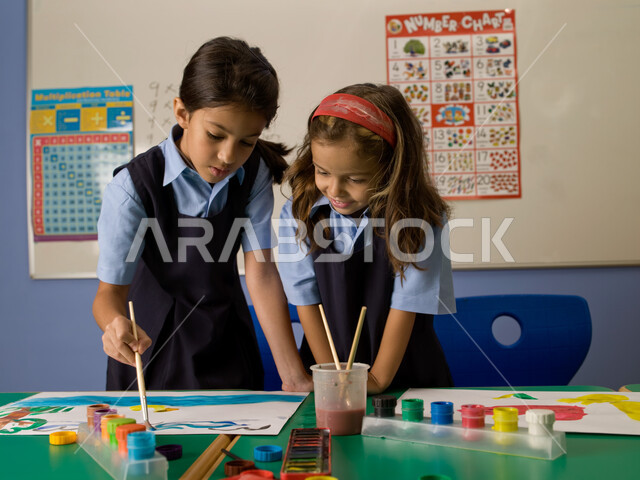أدوات ومعدات التلوين والرسم، التعلم وتنمية المهارات الفنية في المدارس الإماراتية، طالبتان عربيتان خليجيتان إماراتيتان ترتديان الزي المدرسي تتشاركان بتلوين الرسومات بالألوان المائية، الاندماج والتركيز في طلاء الصور، أنشطة ترفيهية جماعية في الفصل الدراسي.