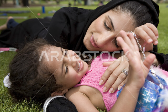 ايماءات طفلة تدل على الأمان و الحب, الاعتناء بالأطفال والاهتمام بهم, مشاعر الأمومة و العطف، صورة مقربة لأم عربية خليجية إماراتية تعانق إبنتها بحنان في الخارج, الاستلقاء على العشب في إحدى الحدائق العامة, مفهوم الأسرة السعيدة