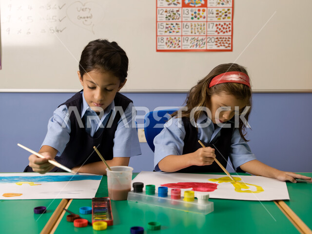 Learning and developing artistic skills in Emirati schools, coloring tools and equipment, two Arab Gulf Emirati students wearing school uniforms painting with watercolors, integration and focus in painting pictures, group recreational activities in the cl