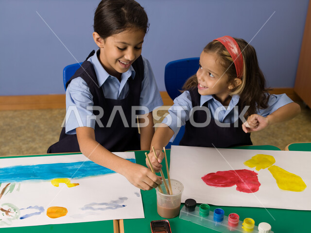 إيماءات تدل على الاندماج والسعادة والاستمتاع في طلاء الصور، التعلم وتنمية المهارات الفنية في المدارس الإماراتية، طالبتان عربيتان خليجيتان إماراتيتان ترتديان الزي المدرسي تقومان بالرسم والتلوين بالألوان المائية، أنشطة ترفيهية جماعية في الفصل الدراسي.