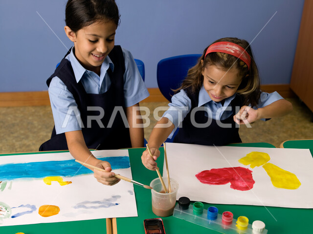 إيماءات تدل على الاندماج والسعادة والاستمتاع في طلاء الصور، التعلم وتنمية المهارات الفنية في المدارس الإماراتية، طالبتان عربيتان خليجيتان إماراتيتان ترتديان الزي المدرسي تقومان بالرسم والتلوين بالألوان المائية، أنشطة ترفيهية جماعية في الفصل الدراسي.
