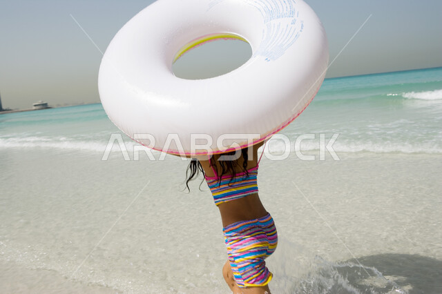 Spending fun times in the sea, happiness and enjoying summer activities, beach resorts in the Emirates, a back image of a young Arab Emirati Gulf girl running on the beach holding a swimming buoy in her hand, entertaining leisure activities on vacation