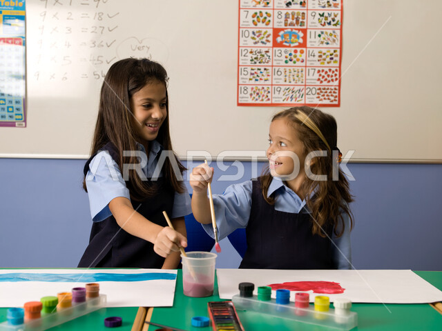 إيماءات تدل على السعادة والاستمتاع بالتعاون والتشارك بالأنشطة الترفيهية الجماعية في الفصل الدراسي، التعلم وتنمية المهارات الفنية في المدارس الإماراتية، طالبتان عربيتان خليجيتان إماراتيتان ترتديان الزي المدرسي تقومان بالرسم والتلوين بالألوان المائية.