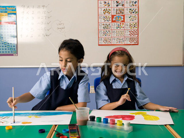 إيماءات تدل على السعادة والاستمتاع بالتعاون والتشارك بالأنشطة الترفيهية الجماعية في الفصل الدراسي، التعلم وتنمية المهارات الفنية في المدارس الإماراتية، طالبتان عربيتان خليجيتان إماراتيتان ترتديان الزي المدرسي تقومان بالرسم والتلوين بالألوان المائية.