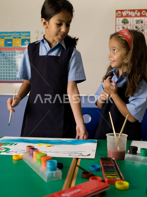 التعلم وتنمية المهارات الفنية في المدارس الإماراتية، إيماءات تدل على السعادة والاستمتاع بالتعاون والتشارك بالأنشطة الترفيهية الجماعية في الفصل الدراسي،  طالبتان عربيتان خليجيتان إماراتيتان ترتديان الزي المدرسي تقومان بالرسم والتلوين بالألوان المائية.