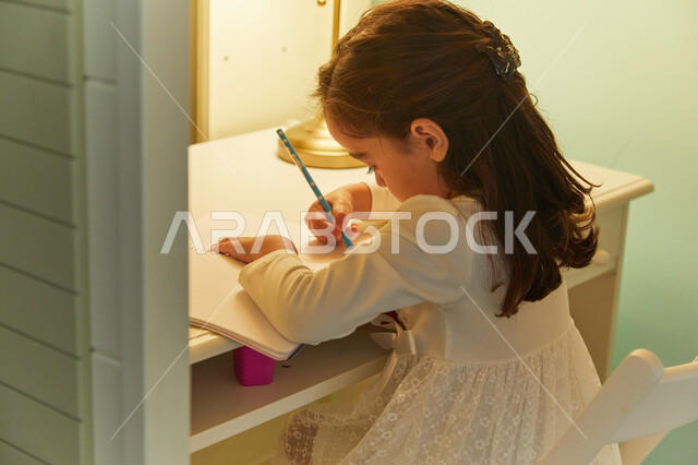 الفصل الدراسي جديد، التعليم والدراسة في المنزل أثناء الحجر الصحي، القيام بتأديه الواجبات المنزليه، صوره من الخلف  لطالبه صغيرة عربيه خليجية اماراتية تجلس على طاولة الدراسة وتقوم بحل الوظائف.