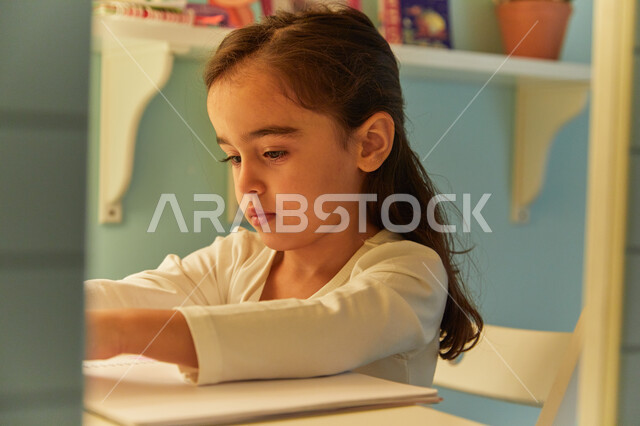 Gestures indicating focus and thinking about the answer, doing homework, a close-up of a young Arab Gulf Emirati student sitting at the study table and doing jobs, the concept of education and studying at home during quarantine.