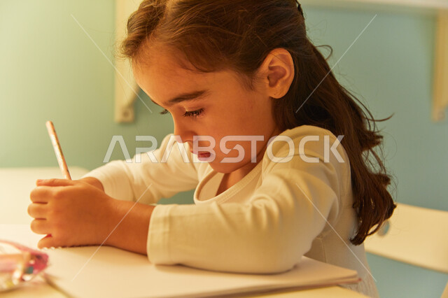 القيام بتأديه الواجبات المنزليه، صوره مقربه لطالبه صغيرة عربيه خليجية اماراتية تجلس على طاولة الدراسة وتقوم بحل الوظائف، ايماءات تدل على التركيز والتفكير بالإجابة، مفهوم التعليم والدراسة في المنزل أثناء الحجر الصحي.