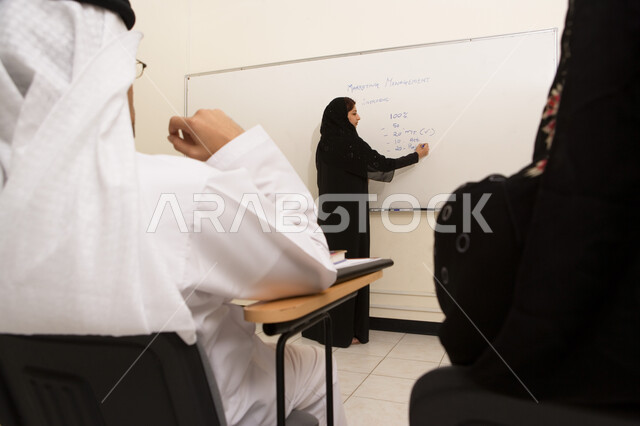 تطوير أساليب التعليم الجامعي في الإمارات، الإنتباه والتركيز والإستماع إلى المحاضرة، صورة مقربة من الخلف لمعلمة جامعية عربية خليجية إماراتية تقوم بتدوين الملاحظات على السبورة أثناء شرح المحاضرة للطلاب، ورش عمل ودورات تدريبية، التعليم الجامعي الإماراتي