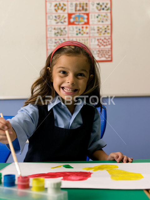 إيماءات تدل على السعادة والاستمتاع بالقيام بالأنشطة الترفيهية في الفصل الدراسي، التعلم وتنمية المهارات الفنية في المدارس الاكاديمية الإماراتية، طالبة عربية خليجية إماراتية ترتدي الزي المدرسي تقوم بالرسم والتلوين بالألوان المائية، النظر إلى الكاميرا.