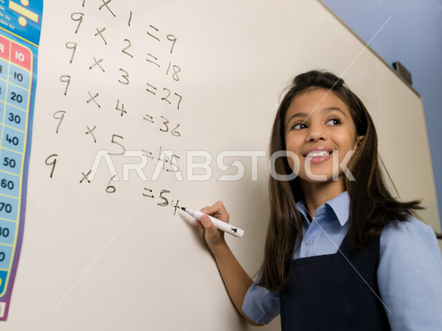 تعلم جدول الضرب والعمليات الحسابية في الرياضيات، جودة التعليم والمدارس في الإمارات العربية المتحدة، صورة مقربة لتلميذة عربية خليجية إماراتية ترتدي الزي المدرسي تقوم بحل التمارين على السبورة، تنمية القدرات الفكرية ومهارات الحساب لطلاب المرحلة الابتدائية.