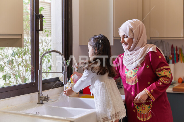 Raising children to love cooperation, to enjoy doing housework with their children, an Arab Gulf Emirati mother standing with her daughter in the kitchen, completing routine tasks at home, helping to clean and wash dishes.