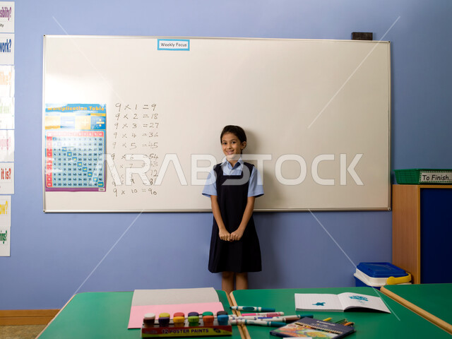  تقوية مهارات الحساب لتلاميذ المرحلة الابتدائية، تعلم جدول الضرب والعمليات الحسابية في الرياضيات، جودة التعليم في مدارس الإمارات العربية المتحدة، تلميذة عربية خليجية إماراتية ترتدي الزي المدرسي تقف بإيماءات الثقة بالحل الصحيح، قرطاسية وأدوات مدرسية.