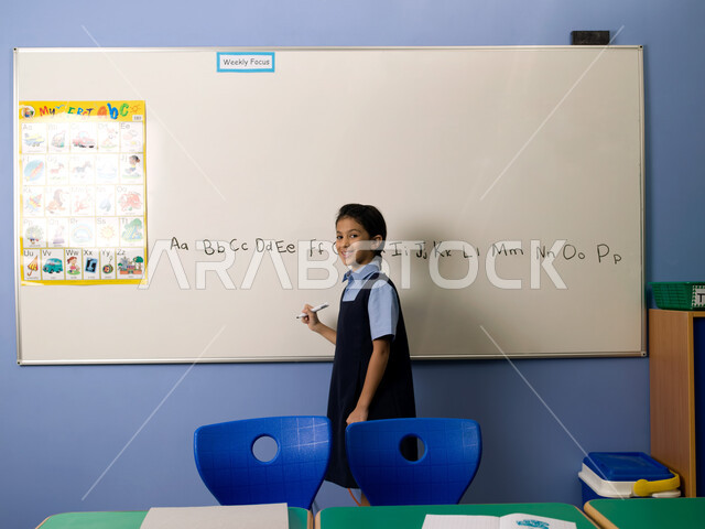 Teaching foreign languages to primary school students, the correct way to draw and write English letters, modern and developed schools in the United Arab Emirates, an Emirati Arab Gulf student wearing a school uniform standing in the school classroom, fac